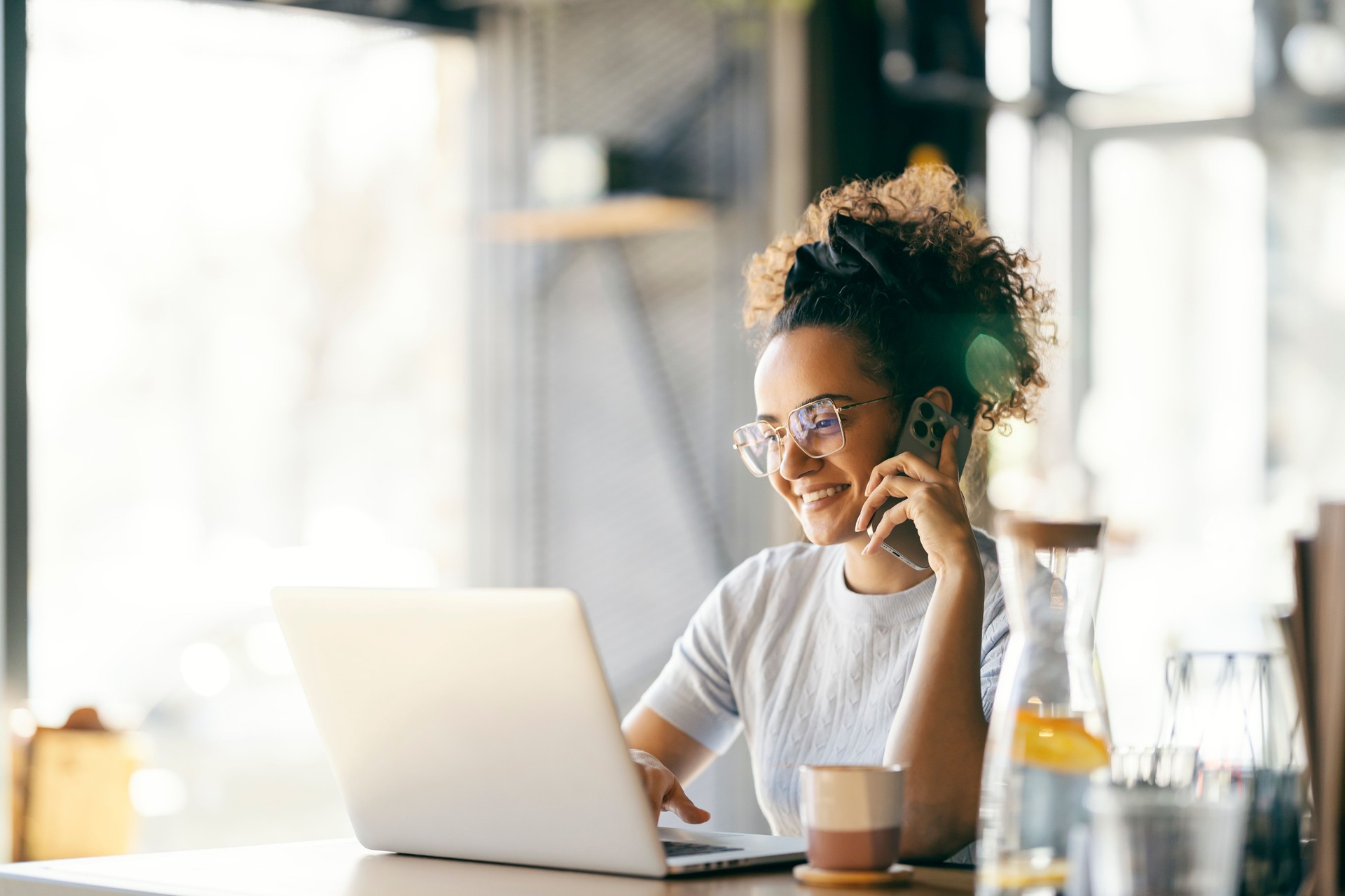 Portrait of young freelancer sitting in cafe, having phone conversation and typing documentation on laptop.